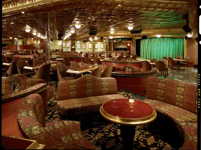 Luxuriöse Lounge der Carnival Conquest. Rote Polstermöbel, Tische, Bühne mit grünen Vorhängen. Goldene Deckenverzierungen und viele Lichter schaffen ein elegantes Ambiente. Keine Personen sichtbar.
