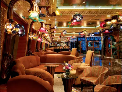 Blick in eine farbenfrohe Lounge der Carnival Conquest. Zahlreiche braune Sofas und Sessel stehen um kleine Tische. Die Decke ist mit leuchtenden, blumenförmigen Glaslampen in vielen Farben dekoriert. Ein glänzender Marmorboden reflektiert das Licht.