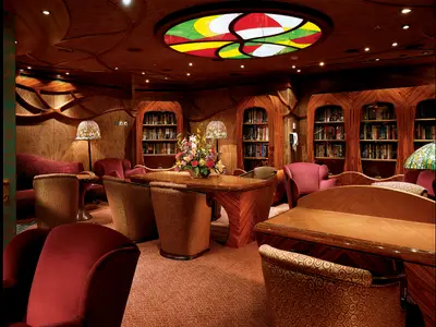 Elegante Schiffsbibliothek mit dunklen Holzregalen voller Bücher. Rote Sessel und gemusterte Stühle um einen Holztisch mit Blumen. Eine bunte Glasdecke spendet warmes Licht.