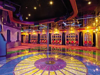 Ein leerer Nachtclub auf der Carnival Conquest mit einem leuchtenden, bunten Glasboden, roten Stühlen entlang der Wände und einer zentralen Stange. Die Decke ist spiegelnd mit Bühnenbeleuchtung und Rohren verziert. Die Wände sind als Ziegelmauerwerk und verputzt mit Laternen gestaltet.