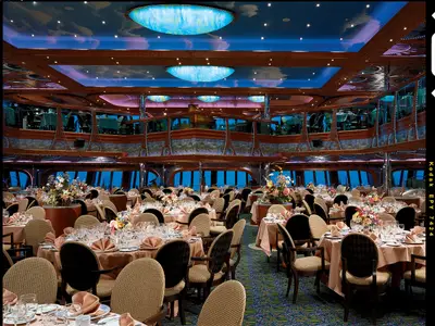 Großer, luxuriöser Speisesaal der Carnival Conquest, festlich gedeckte Tische, üppige Blumenarrangements und goldene Verzierungen. Blaue, bewölkte Decke leuchtet dezent.