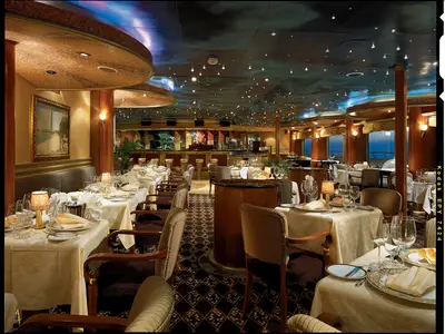 Innenansicht eines eleganten Restaurants auf der Carnival Conquest. Zahlreiche Tische sind weiß eingedeckt mit Gläsern, Tellern und Servietten. Im Hintergrund ist eine Bar zu sehen. Große Fenster geben den Blick auf das dunkle Meer frei. Die Decke ist mit Sternlichtern verziert.