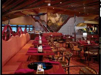 Ein großes, elegantes Restaurant auf der Carnival Conquest mit zahlreichen roten Tischen, Stühlen und Sitzbänken. Eine auffällige Treppe führt zu einer oberen Ebene mit einer künstlerischen Mosaikwand.
