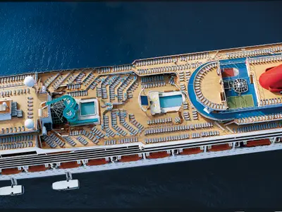 Vogelperspektive auf das belebte Sonnendeck der Carnival Conquest mit mehreren Pools, einer blauen Wasserrutsche, zahlreichen Liegestühlen und einem Sportfeld, umgeben von blauem Meer.