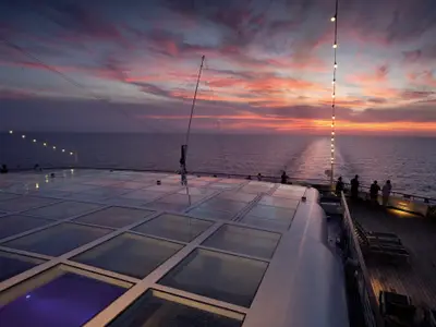 Blick von oben auf das Sonnendeck der Carnival Conquest bei Sonnenuntergang. Menschen stehen an der Reling und schauen aufs weite Meer, der Himmel ist in leuchtenden Orange- und Rottönen gefärbt. Eine Lichtinstallation aus Glühbirnen hängt herab. Ein Bereich des Decks ist mit Glasplatten bedeckt.