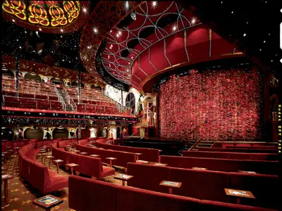 Ein prachtvolles Theater auf der Carnival Conquest mit mehreren Ebenen roter Samtsitze, einer kunstvollen Bühne mit schwerem rotem Vorhang und einer dunklen Decke, die mit funkelnden Lichtern wie Sterne beleuchtet ist.