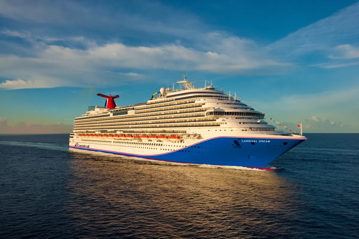 Die Carnival Dream, das blau-weiße Schiff mit markantem roten Schornstein, fährt auf ruhigem, blauem Meer unter leicht bewölktem Himmel. Der Schiffsname ist am Bug sichtbar.