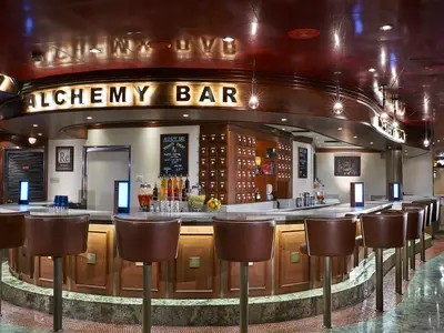 Blick in die stilvolle Alchemy Bar der Carnival Dream. Eine lange Theke mit braunen Lederbarhockern, dahinter Regale und beleuchtete Schilder. Die Decke ist rot glaenzend, der Boden mit Fliesenmosaik.