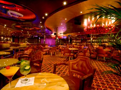 Eine aufwendig dekorierte Bar-Lounge auf der Carnival Dream mit rotem Teppich, dunklen Holzmöbeln und zwei Cocktails im Vordergrund. Rote Kronleuchter und indirekte Beleuchtung schaffen eine lebhafte Atmosphäre.