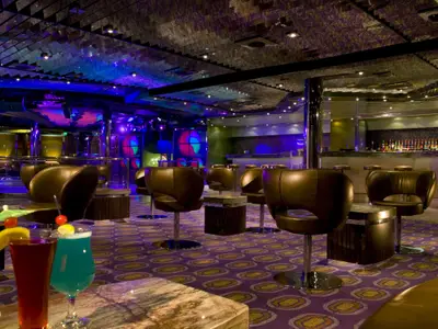 Innenansicht einer modernen Bar auf der Carnival Dream mit lila Teppich, braunen Barhockern und farbiger LED-Beleuchtung an der Decke und den Wänden. Im Hintergrund ist eine gut sortierte Bar zu sehen und vorne zwei Cocktails.