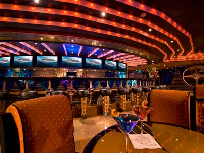 Lebhafte Bar-Lounge auf der Carnival Dream mit farbigen Lichtstreifen an der Decke. Mehrere Bildschirme zeigen Schiffe. Im Vordergrund stehen zwei bunte Cocktails auf einem Tisch.