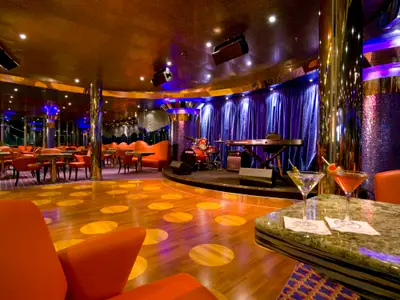 Blick in eine lebhafte Bar und Lounge auf der Carnival Dream mit einer beleuchteten Bühne. Rote Sitze, Holzboden mit Kreismustern, und blaue Vorhänge sind zu sehen. Im Vordergrund stehen zwei Cocktails auf einem Tresen.