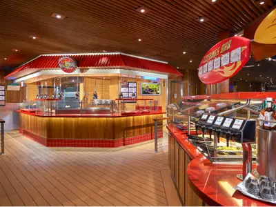 Ein lebhaftes Guy's Burger Joint und Toppings Bar auf der Carnival Dream. Die Theken sind rot mit Holzverkleidung, es gibt verschiedene Saucen und Beläge, und ein TV-Bildschirm zeigt Guy Fieri.