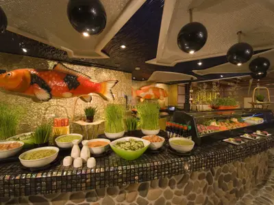 Übersicht eines asiatischen Buffets an Bord der Carnival Dream. Eine Auswahl an Sushi, Salaten und Soßen ist auf einem langen Tresen mit glänzenden Mosaikfliesen angerichtet. Große, orangefarbene Koi-Fisch-Skulpturen schmücken die goldene Wand im Hintergrund.