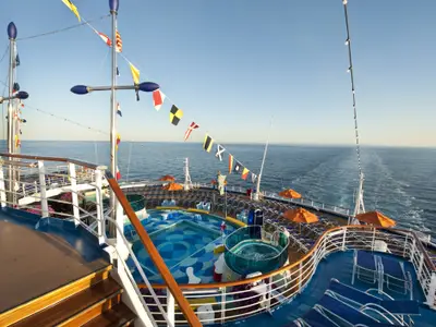 Blick über das Oberdeck der Carnival Dream mit einem großen Pool und einem Whirlpool. Zahlreiche leere Liegestühle unter orangefarbenen Sonnenschirmen säumen den Bereich. Im Hintergrund ist das weite blaue Meer unter klarem Himmel zu sehen. Signalflaggen wehen an den Masten.
