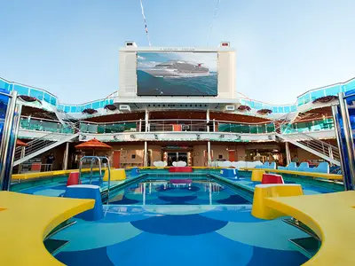 Großer Außenpoolbereich an Deck der Carnival Dream. Bunter Pool, umgeben von Sitzen. Mehrere Ebenen mit Glasbalkonen und ein großer Bildschirm mit Schiffsansicht.