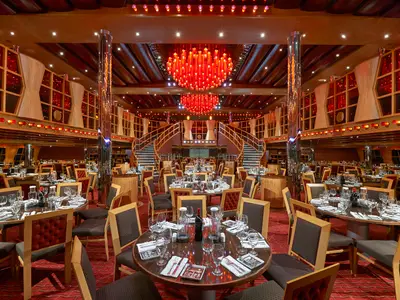 Großes, opulentes Restaurant an Bord der Carnival Dream mit roten Kronleuchtern und doppelt geschwungener Treppe. Viele gedeckte Tische auf rotem Teppich.