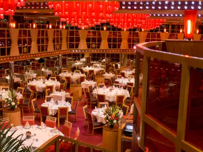 Innenansicht eines mehrstöckigen Restaurants auf der Carnival Dream. Zahlreiche runde und eckige Tische sind mit weißen Tischtüchern und Gedecken vorbereitet. Der Raum ist in Rottönen gehalten, mit roten Lampen an der Decke und roten Teppichen. Eine Balustrade ist im Vordergrund zu sehen.