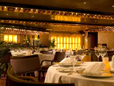 Blick in ein luxuriöses Restaurant mit mehreren weiß gedeckten Tischen, eleganten Stühlen, glänzenden Gläsern und Besteck. Goldene Beleuchtung und Wandverkleidungen schaffen eine festliche Atmosphäre. Eine grüne Pflanze steht links im Vordergrund.