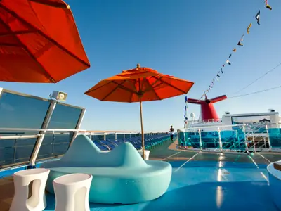 Sonnendeck der Carnival Dream mit orangefarbenen Sonnenschirmen, blauer Lounge und Hockern. Im Hintergrund Person, Liegestühle, Meer und roter Schornstein unter blauem Himmel.