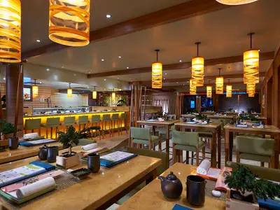 Ein stilvolles asiatisches Spezialitätenrestaurant an Bord der Carnival Dream, mit einer langen beleuchteten Sushi-Bar, Tischen mit grünen Stühlen und warmen Holzdetails. Ohne Personen.