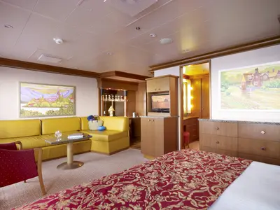 Helle, geräumige Suite an Bord der Carnival Dream mit gelber Ledercouch, Esstisch, Bartresen, TV und Kingsize-Bett mit roter Decke. Ein Bad ist durch einen offenen Durchgang sichtbar.