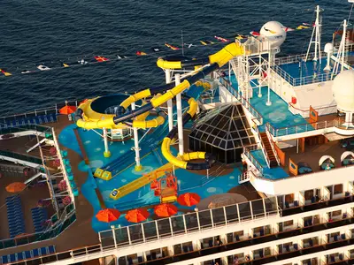 Vogelperspektive auf das Oberdeck der Carnival Dream. Große gelbe und dunkelblaue Wasserrutschen schlängeln sich über einen hellblauen Poolbereich. Liegestühle, Sonnenschirme und Kabinenbalkone sind ebenfalls sichtbar. Das tiefe Blau des Meeres umgibt das Schiff.