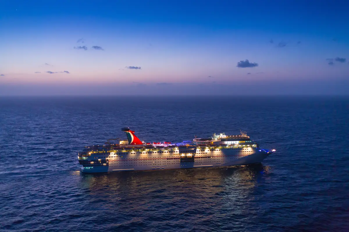Eine große, hell beleuchtete Carnival Elation mit rotem Schornstein fährt bei Dämmerung auf dem dunklen Meer, unter einem violett-blauen Himmel.
