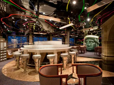 Thematische New York Bar auf der Carnival Elation mit Piano-Bar, Barhockern, Replik des Kopfes der Freiheitsstatue und US-Navy-Luftschiff. Stylisierte Skyline hinter den Fenstern.