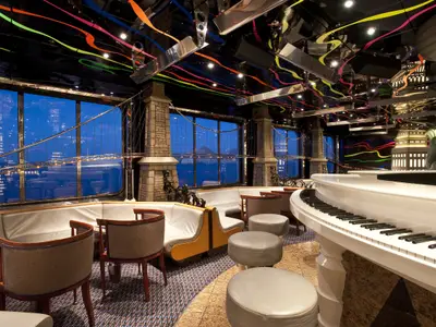 Elegante Bar-Lounge auf der Carnival Elation mit weißem Flügel und bequemen Sitzgelegenheiten. Große Fenster zeigen eine stilisierte New Yorker Skyline. Die Decke ist mit bunten Neonbändern und reflektierenden Paneelen verziert, was eine lebhafte Atmosphäre schafft. Eine Statue im Liberty-Stil ist im Hintergrund zu sehen.