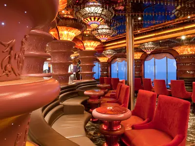 Opulente Bar-Lounge auf der Carnival Elation. Rote Samtsessel und beige Sofas. Der Raum ist reich verziert mit kunstvollen Säulen, bunten Glaslampen und großen Fenstern mit Meerblick.