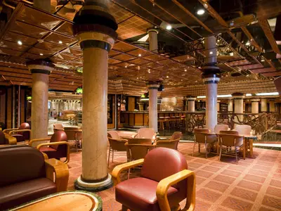 Blick in eine weitläufige Bar-Lounge auf der Carnival Elation. Die Szene zeigt helle Marmorsäulen, eine gemusterte Decke mit Spiegelflächen, diverse Sitzgelegenheiten in Rottönen und eine lange Bar im Hintergrund. Der Boden ist mit einem warmfarbigen, gemusterten Teppich ausgelegt. Keine Personen sind sichtbar.