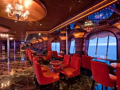 Eine opulent eingerichtete Bar-Lounge auf der Carnival Elation mit bequemen roten Sesseln und kleinen Tischen. Große Fenster bieten einen weiten Blick auf das offene Meer. Die Decke ist mit dekorativen Lichtern und Spiegeln verziert. Der Boden ist glänzend gefliest.