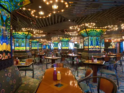 Innenansicht eines farbenprächtigen Buffetrestaurants auf der Carnival Elation. Die Tische sind gedeckt, die Säulen sind mit Pfau-Motiven in Buntglasoptik verziert. Warme Lichtinstallationen an der Decke schaffen eine gemütliche Atmosphäre.