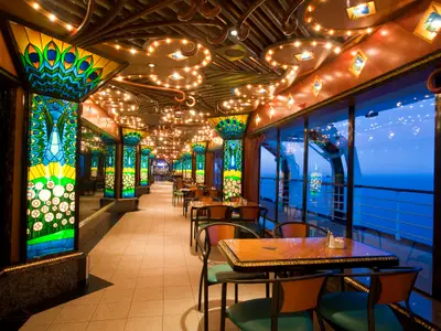 Elegantes Restaurant der Carnival Elation. Wände mit farbenfrohen Buntglasfenstern (Pfauenmotive). Meerblick durch große Fenster. Viele Tische und Stühle. Decke kunstvoll beleuchtet.