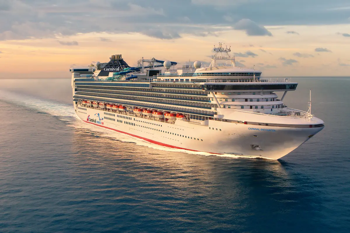 Die Carnival Encounter, ein großes weißes Schiff mit farbigen Logos, fährt bei Sonnenuntergang auf dem ruhigen, blauen Meer. Bug nach rechts, Heck nach links mit einer deutlichen Wellenspur. Der Himmel zeigt warme Orange- und Gelbtöne mit leichten Wolken.