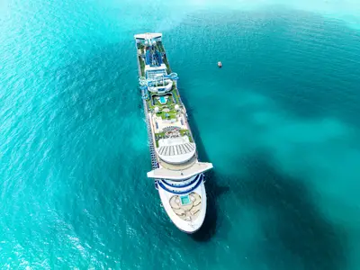 Luftaufnahme der Carnival Encounter auf türkisfarbenem Meer. Auf dem Sonnendeck sind mehrere Wasserrutschen, Pools, Grünflächen und Liegestühle zu sehen. Ein kleines Beiboot schwimmt rechts neben dem Schiff.