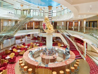 Großes Atrium der luxuriösen Carnival Firenze mit Bar. Goldene Löwenstatue, rote Teppiche, elegante Sitzgruppen, geschwungene Treppen.