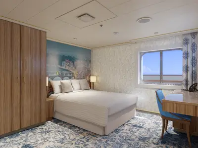 Gerenderte Außenkabine auf der Carnival Firenze mit Doppelbett, hellem Holzschrank und blau gemusterter Tapete. Ein Schreibtisch mit Stuhl und ein maritimes Fenster mit Meerblick sind ebenfalls sichtbar.