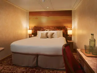 Ein warm beleuchtetes Schlafzimmer auf der Carnival Firenze mit einem großen weißen Doppelbett und zwei bequemen Kissen. Eine Wand ist mit einem malerischen Stadtbild bei Sonnenuntergang dekoriert. Auf Nachttischen stehen Lampen und ein Schreibtisch mit Getränken ist sichtbar.