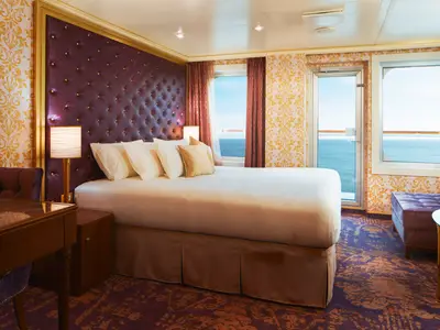 Eine luxuriöse Balkonkabine an Bord der Carnival Firenze. Ein großes Bett mit lila Kopfteil dominiert den Raum. Fenster und Balkontür bieten weiten Meerblick. Elegantes Dekor mit gold-violetten Mustern.