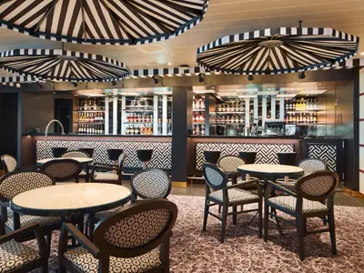Stilvolle Bar-Lounge auf der Carnival Firenze. Gemusterte Stühle an Tischen, gut bestückte Bar. Schwarz-weiß gestreifte Decken-Schirme.