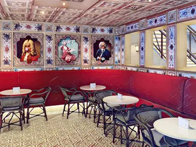 Eine moderne Bar-Lounge auf der Carnival Firenze mit langer roter Sitzbank und kleinen Tischen. Die Wände sind mit humorvoll modernisierten klassischen Porträts dekoriert. Die Decke ist reich gemustert.