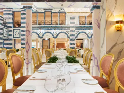 Innenansicht eines luxuriösen Restaurants auf der Carnival Firenze. Die Wände sind mit weißem und blau gestreiftem Marmor verkleidet, unterbrochen von Bögen und Balkonen. Im Vordergrund stehen elegant gedeckte Tische mit weißen Tischtüchern und goldenen Stühlen. Warme Lichtinstallationen sorgen für eine stimmungsvolle Atmosphäre.