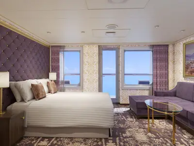 Eine luxuriöse, modern eingerichtete Suite der Carnival Firenze mit einem großen Bett, einer lila Samt-Sitzecke und bodentiefen Fenstern zum Meer. Die Wände sind mit goldenem Damastmuster und einer gepolsterten Kopfwand versehen.