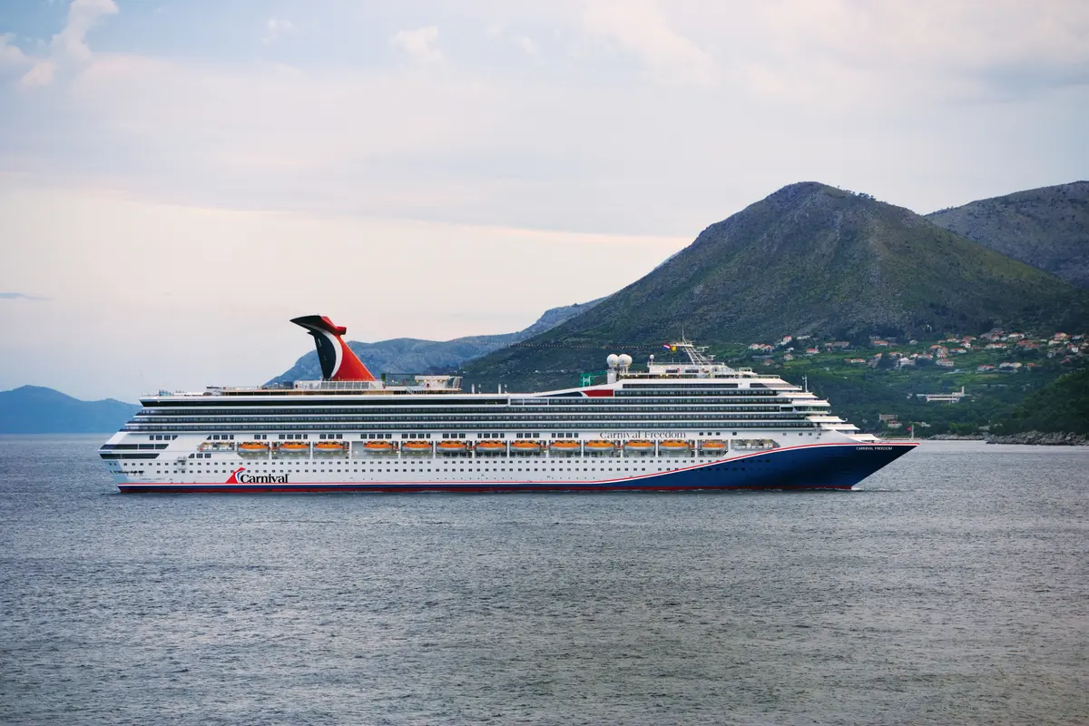 Die große Carnival Freedom mit rotem Schornstein und blauer Rumpfseite fährt bei Tageslicht auf dem Meer. Im Hintergrund sind grüne Berge und eine Küstenstadt zu sehen.