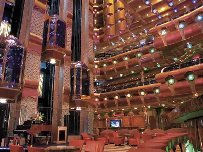 Ein aufwendig dekoriertes Atrium der Carnival Freedom mit mehreren Stockwerken, glänzenden Glasaufzügen, einer Bar mit Sitzgelegenheiten und einem Flügel mit Blumenschmuck. Zahlreiche Lichtinstallationen schaffen eine warme Atmosphäre.