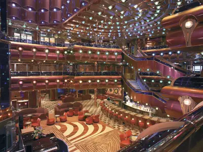Blick von oben in das beeindruckende, mehrstufiges Atrium der Carnival Freedom im Art-déco-Stil. Rote Sitzmöbel, eine geschwungene Bar und prunkvolle Kupferverkleidungen prägen den Raum. Viele glitzernde Lichter erleuchten die Decke.