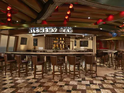 Eine moderne Bar mit braunen Barhockern und einer langgezogenen Theke. Im Hintergrund ist durch Fenster das Meer zu sehen. Die Decke ist kunstvoll mit roten Lichtern gestaltet. Das Schild "Alchemy Bar" leuchtet.