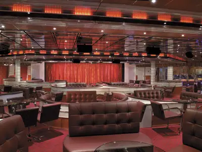 Geräumige Lounge auf der Carnival Freedom mit dunkelroten Ledersofas und Glastischen. Im Hintergrund eine Bühne mit roten Vorhängen. Decke mit orangefarbenen Lichtleisten. Roter Teppich. Ohne Personen.